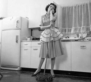 1950s Bored Woman Housewife Wearing Apron Leaning On Mop On Kitchen Floor